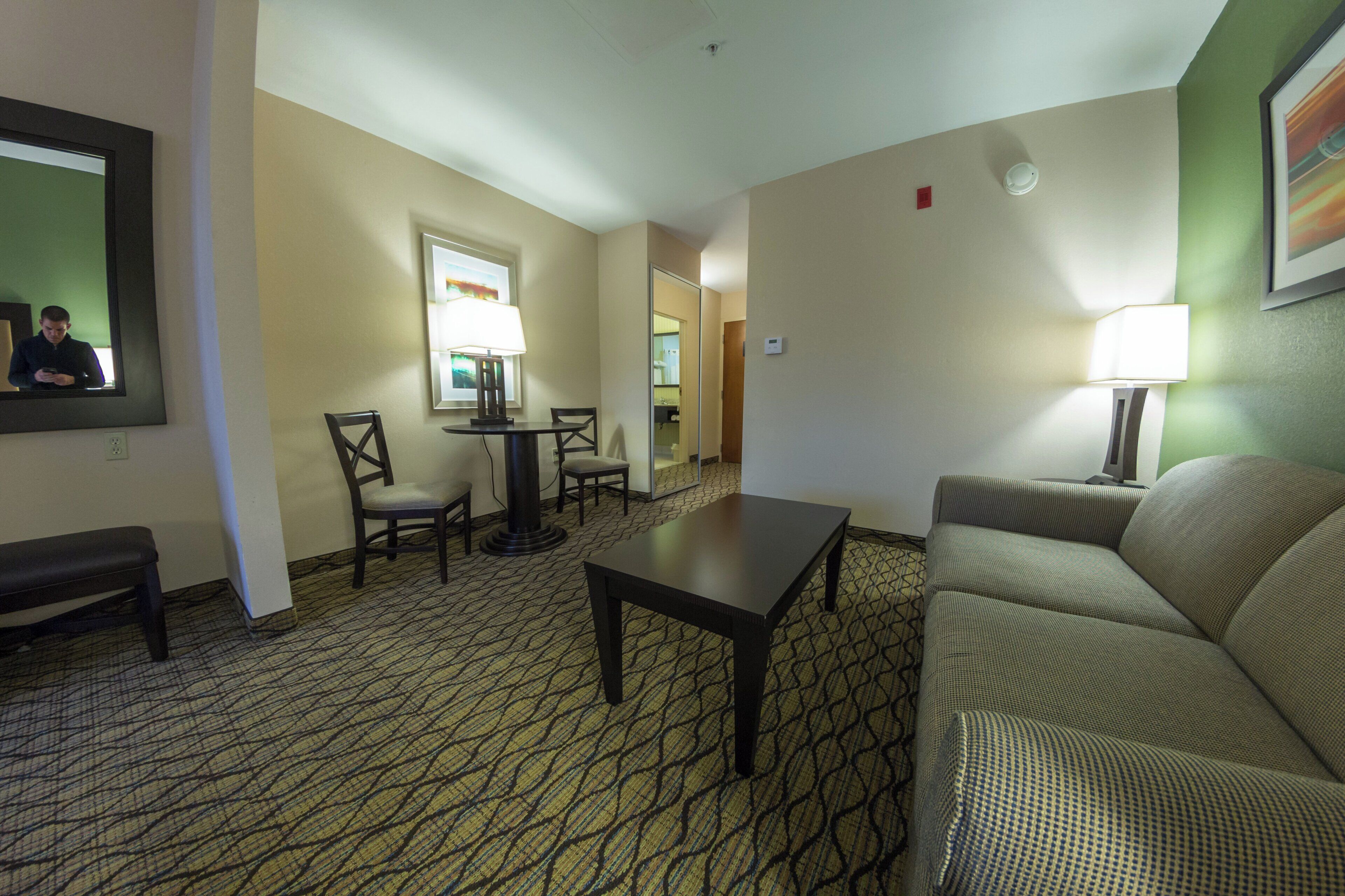 A hotel room with a sofa, a small table, a chair, a lamp, and a mirror.