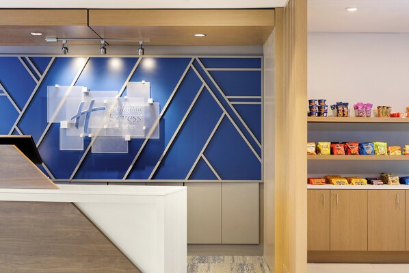 A hotel reception area with a modern design, featuring a blue geometric wall pattern, a reception desk, and a snack shelf with various packaged goods. - Holiday Inn Express Hotel & Suites Winchester by IHG (Winchester)