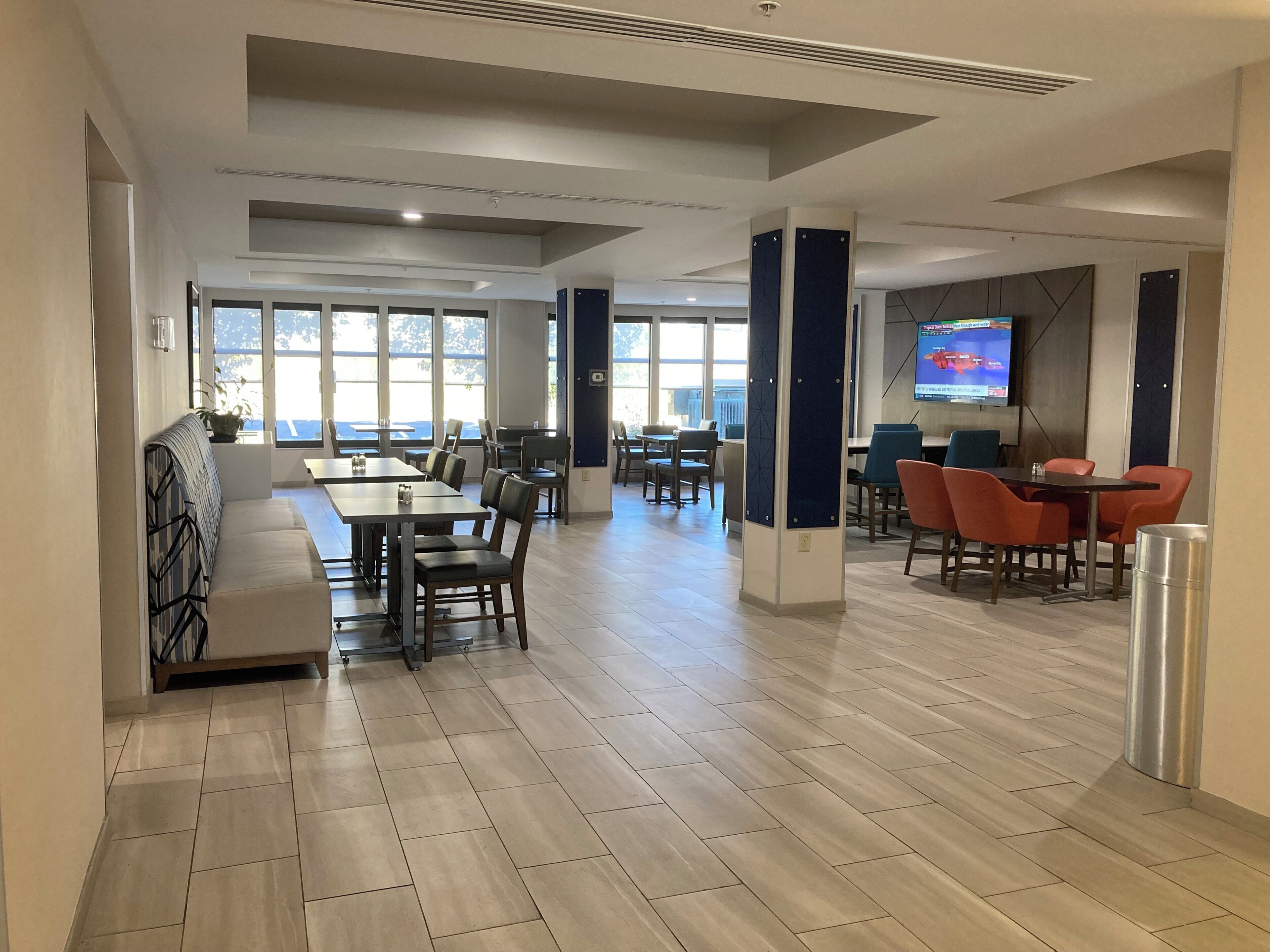 A modern dining area with tables and chairs, large windows, and a flat-screen TV mounted on the wall.