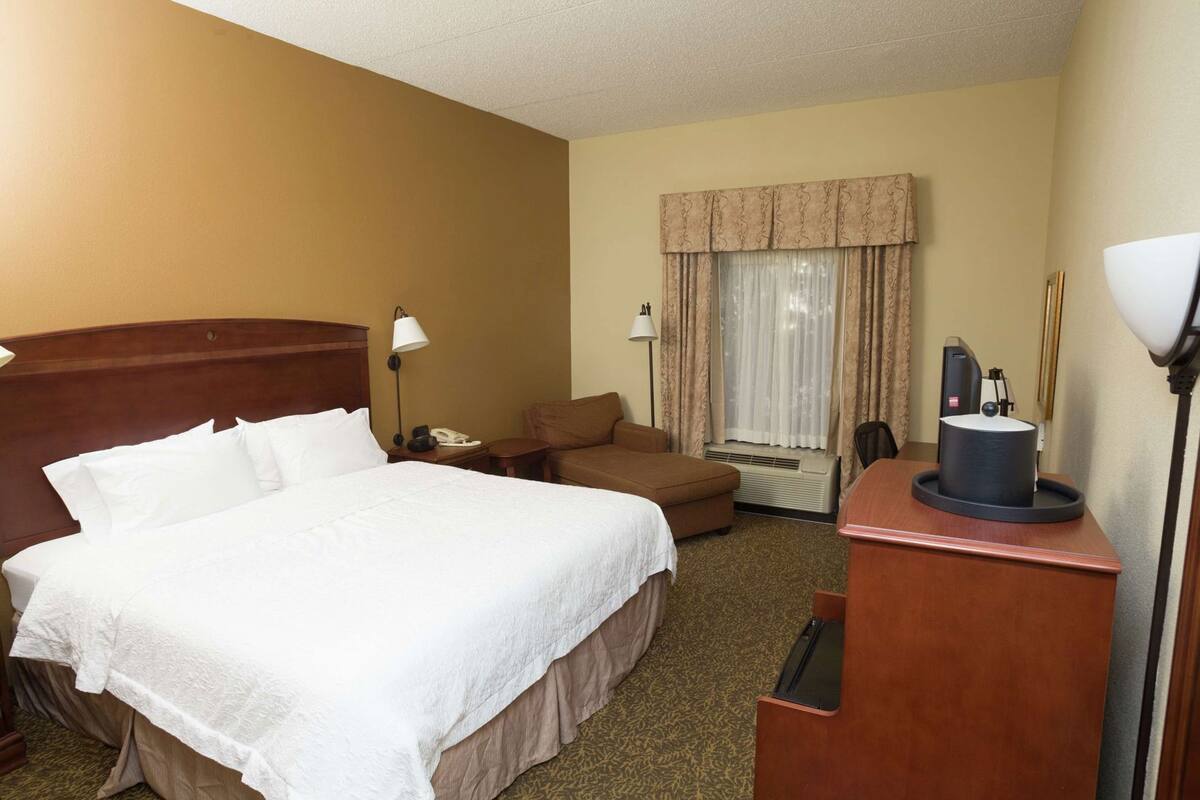 Una habitación de hotel con una cama grande, un escritorio con lámpara, una silla, un sofá y una ventana con cortinas.