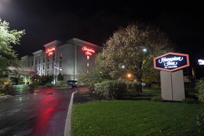 A Hampton Inn hotel at night with illuminated signage, a well-maintained lawn, and a parking area.