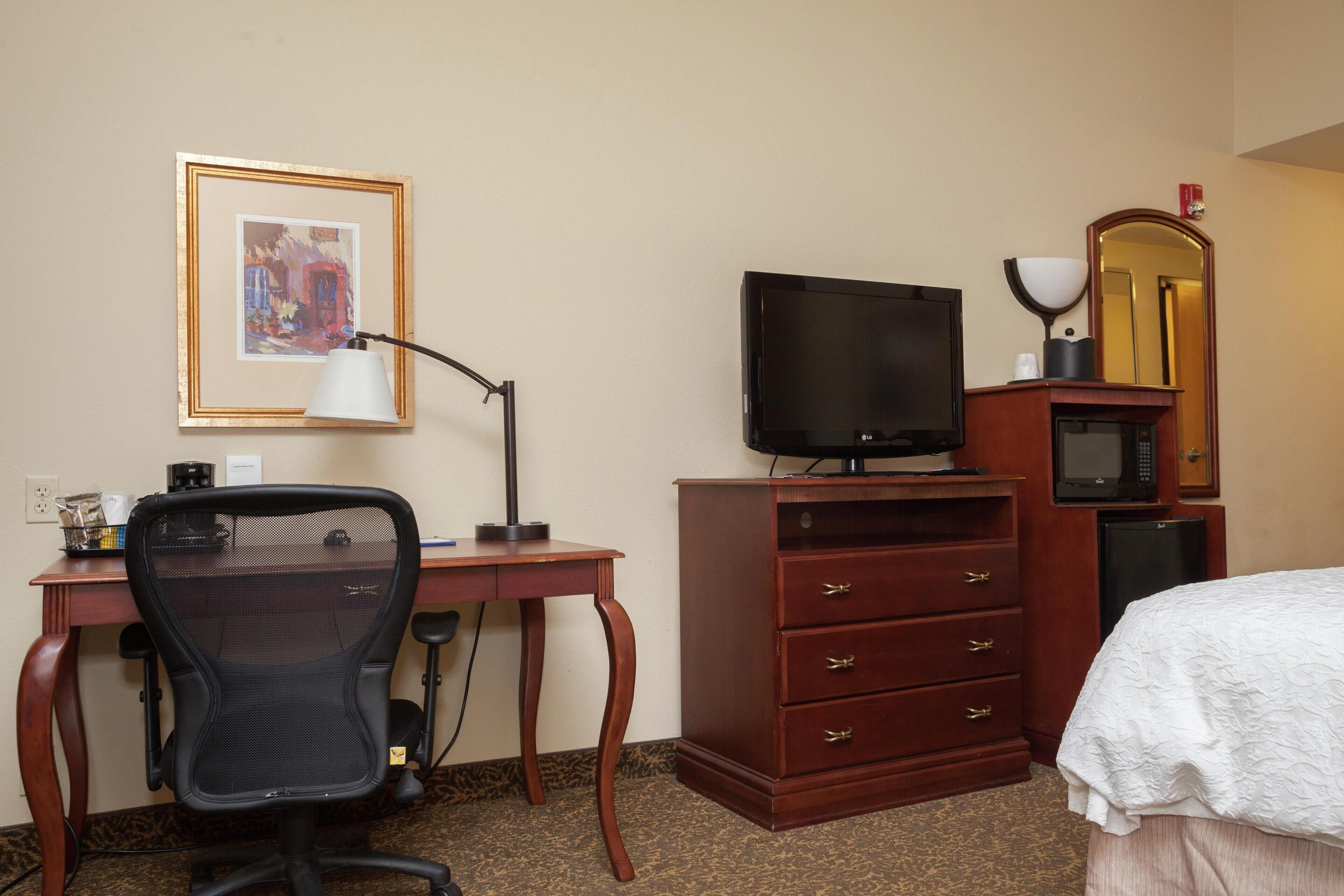 A hotel room with a wooden desk, a black office chair, a television, a dresser, and a bed with a white bedspread.