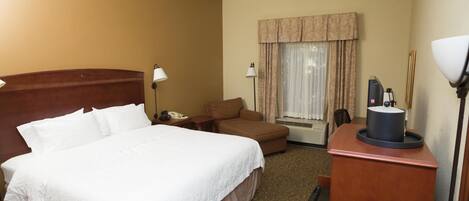 A hotel room with a large bed, a desk with a lamp, a chair, a desk lamp, a telephone, and a window with curtains.