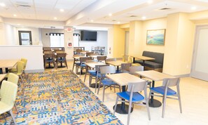 A dining area with multiple tables and chairs, a patterned carpet, and a television screen on the wall.