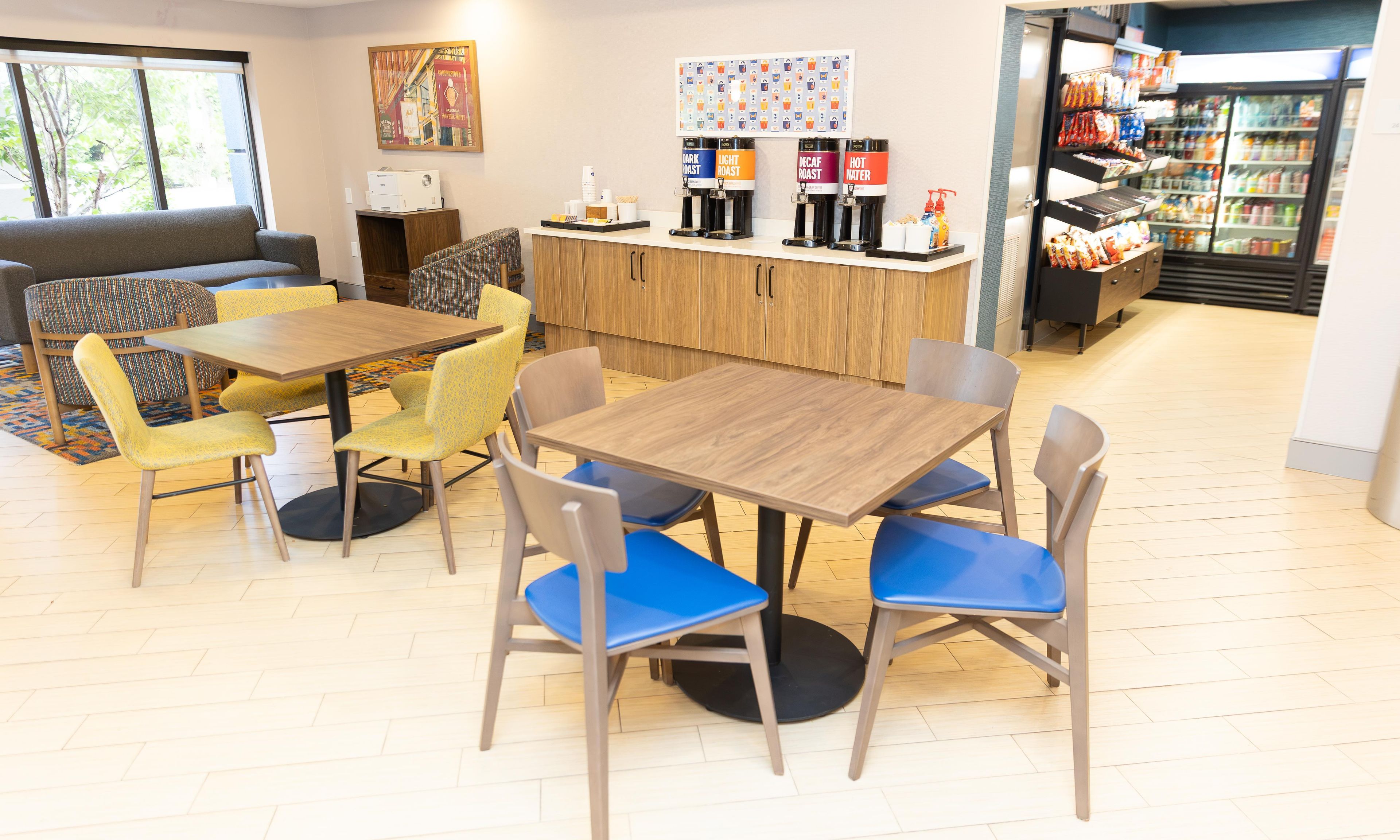 A cafe with wooden tables and chairs, a coffee station, and a refrigerator stocked with snacks.