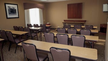 A conference room with long tables and chairs, a large window with curtains, a framed picture on the wall, and a television mounted on the wall.
