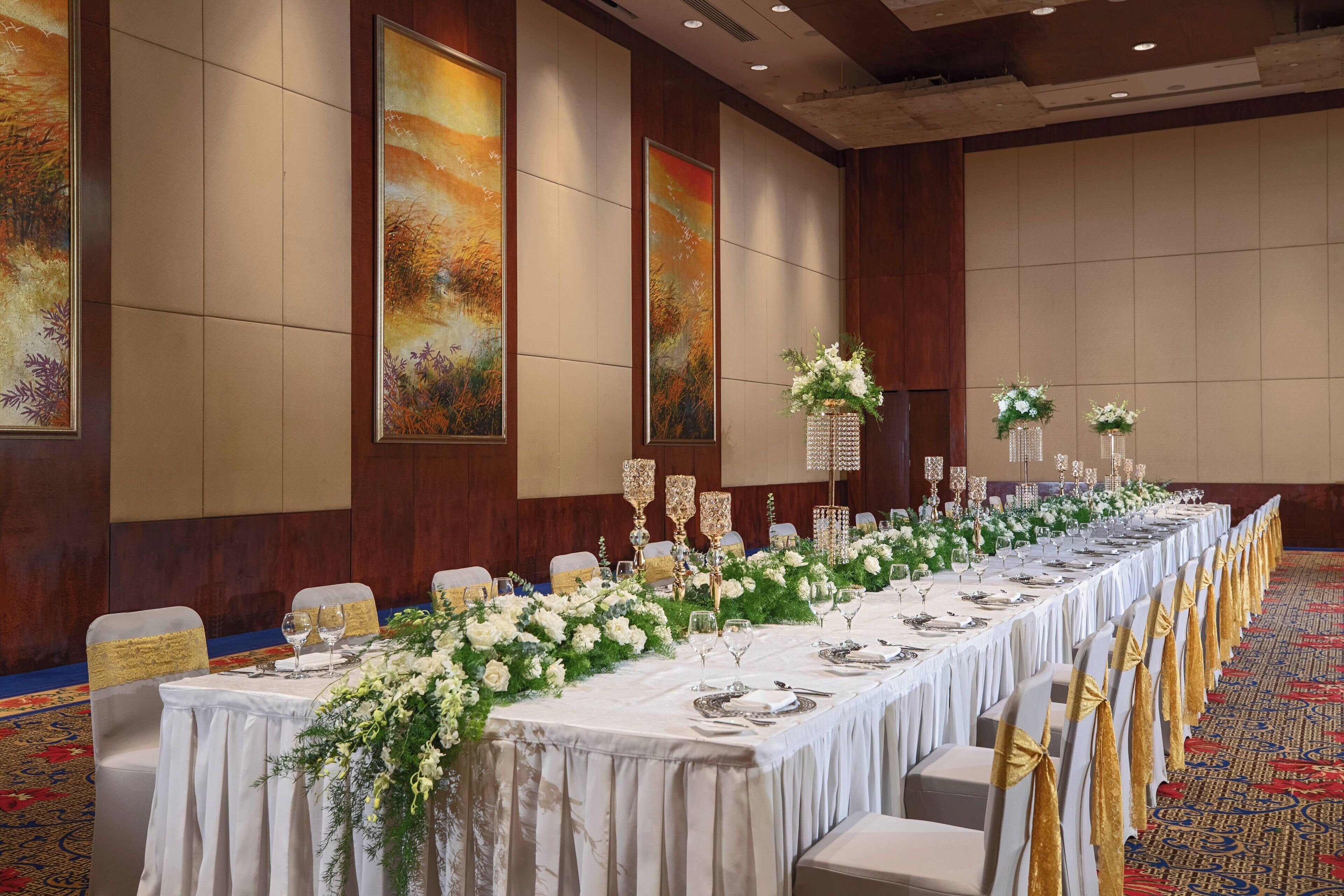 Een balzaal met een lange tafel gedekt voor een formeel evenement, met stoelen met gouden linten, en grote schilderijen aan de muren.
