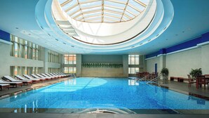 A large indoor swimming pool with a skylight ceiling, sun loungers along the sides, and a seating area with tables and chairs.