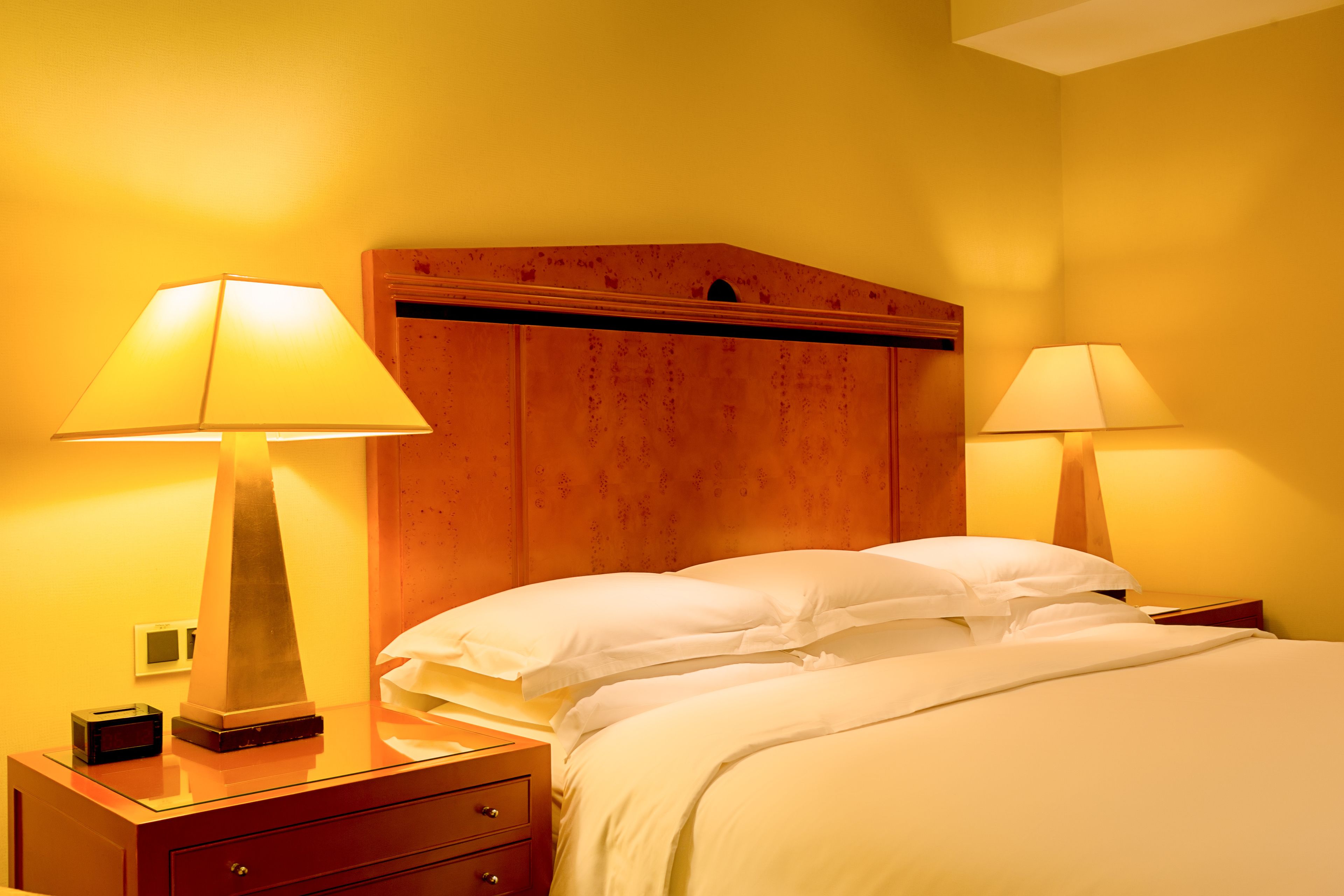 A hotel room with a wooden headboard, two bedside lamps, a bed with white linens, and a nightstand with a lamp and a small box.