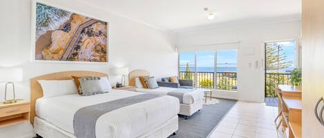 A modern hotel room with two beds, a balcony, and a large window offering a view of the sea.