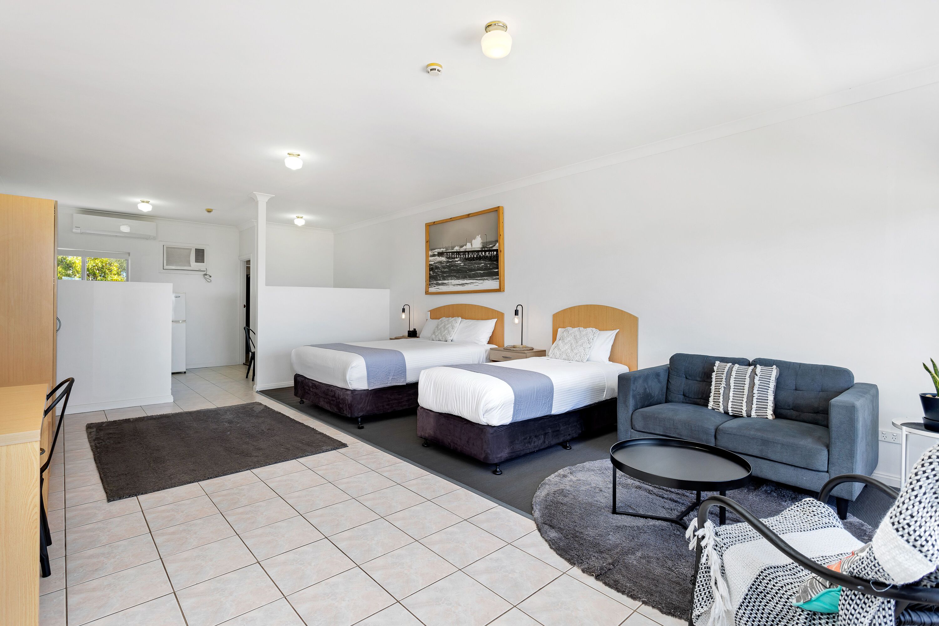 A modern hotel room with two beds, a sofa, a dining area, and a kitchen.