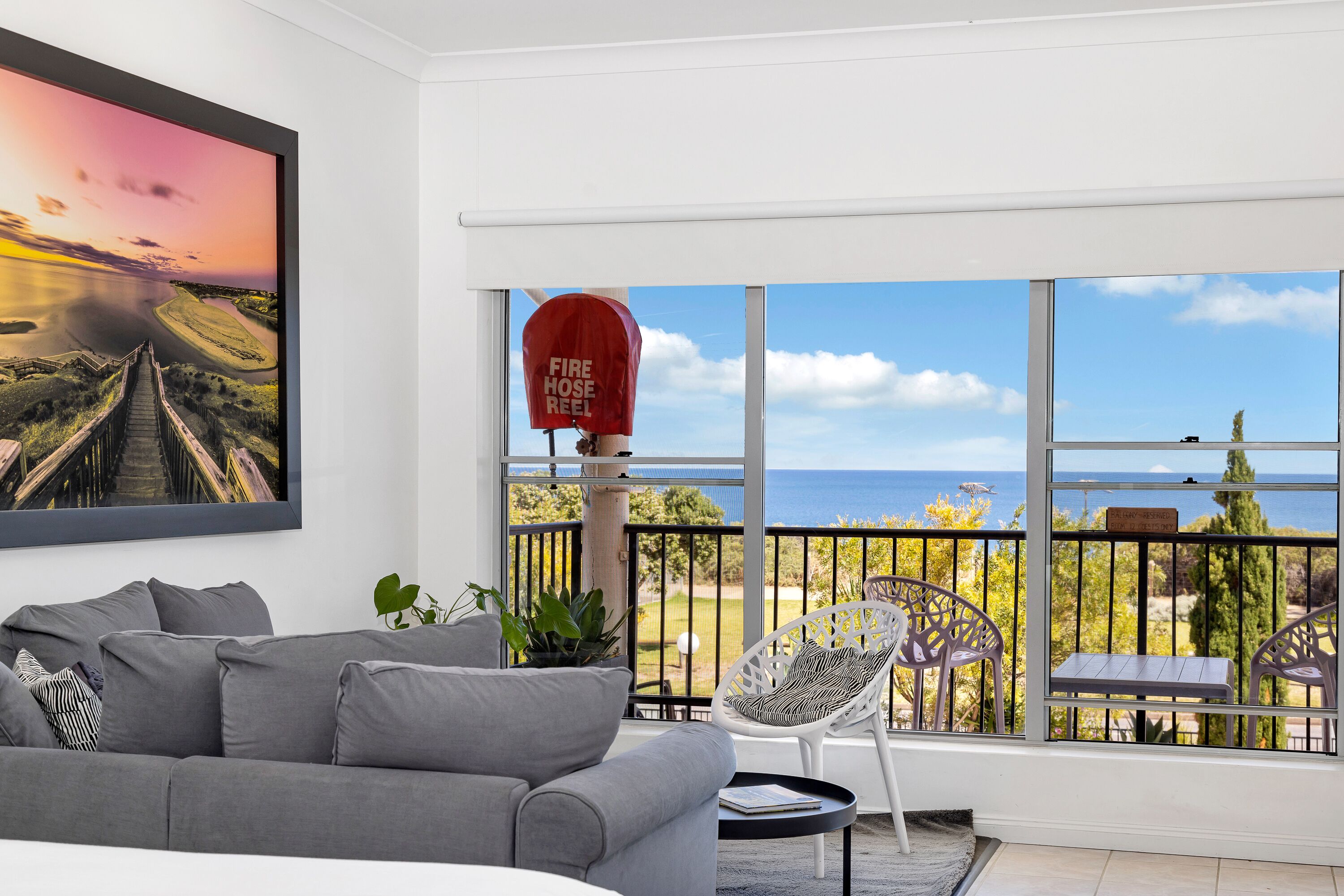 Una sala de estar moderna con un sofá gris, un cuadro con paisaje, un carretel rojo para manguera contra incendios y un balcón con vista al mar.