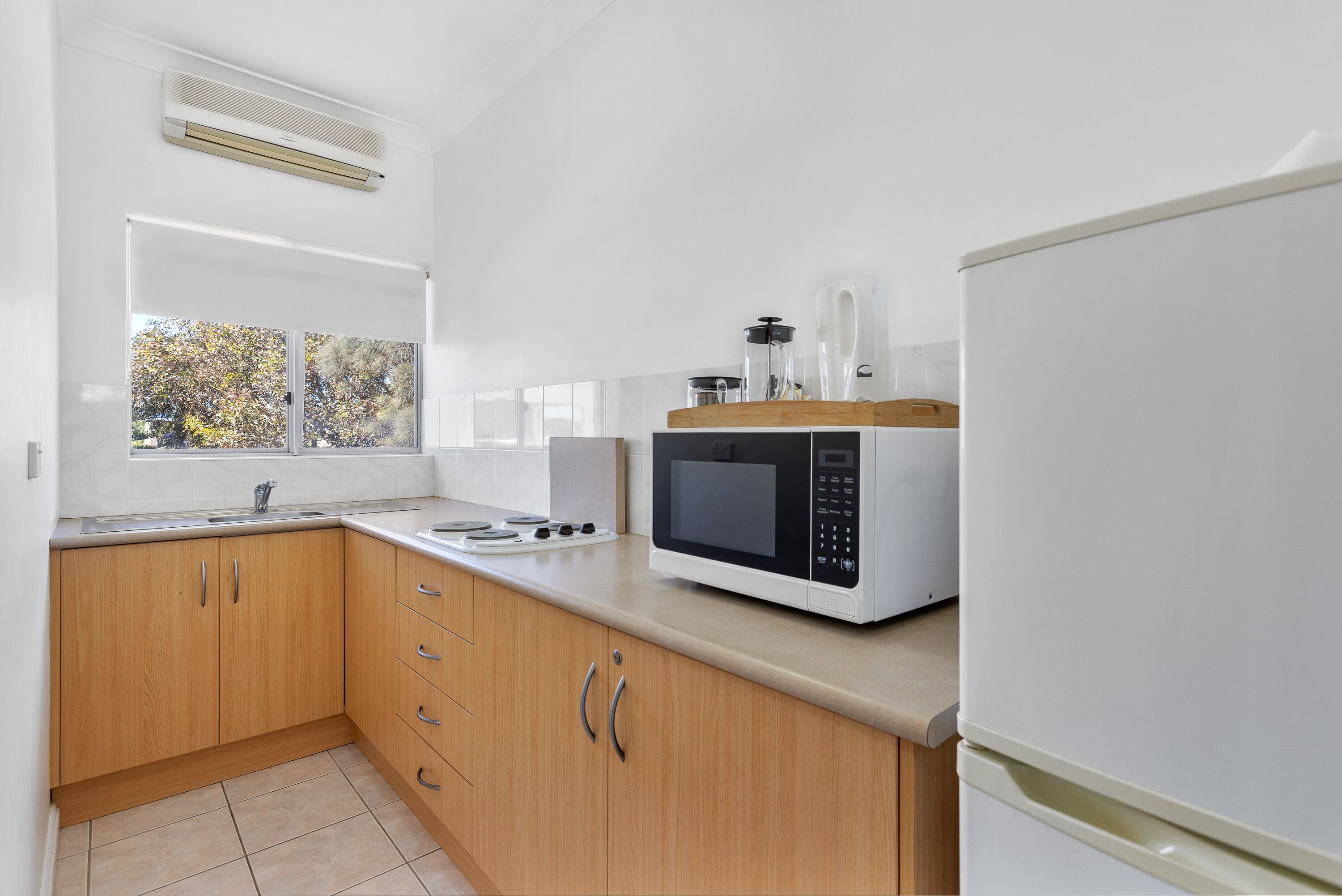 A modern kitchen with wooden cabinets, a white countertop, a built-in microwave, and a window with a view of trees.