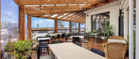 Un espace extérieur aménagé pour les repas, avec des tables et des chaises en bois, des plantes en pot et une vue sur l’océan.