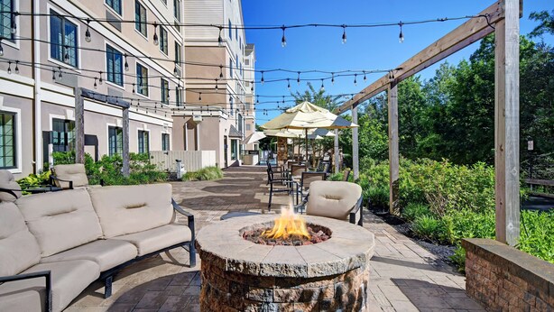 A fire pit in the center of a patio area with outdoor seating and a covered seating area in the background. - Homewood Suites by Hilton Eatontown (Eatontown)