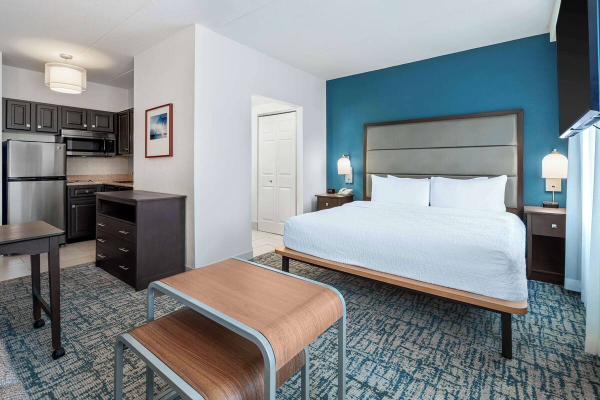 A hotel room with a bed, a desk, a chair, a kitchenette, and a bathroom door.