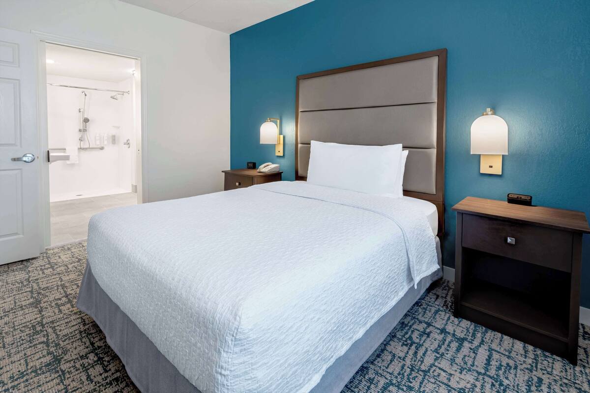 A hotel room with a large bed, two bedside tables, a nightstand, a wall-mounted lamp, and a bathroom visible through an open door.