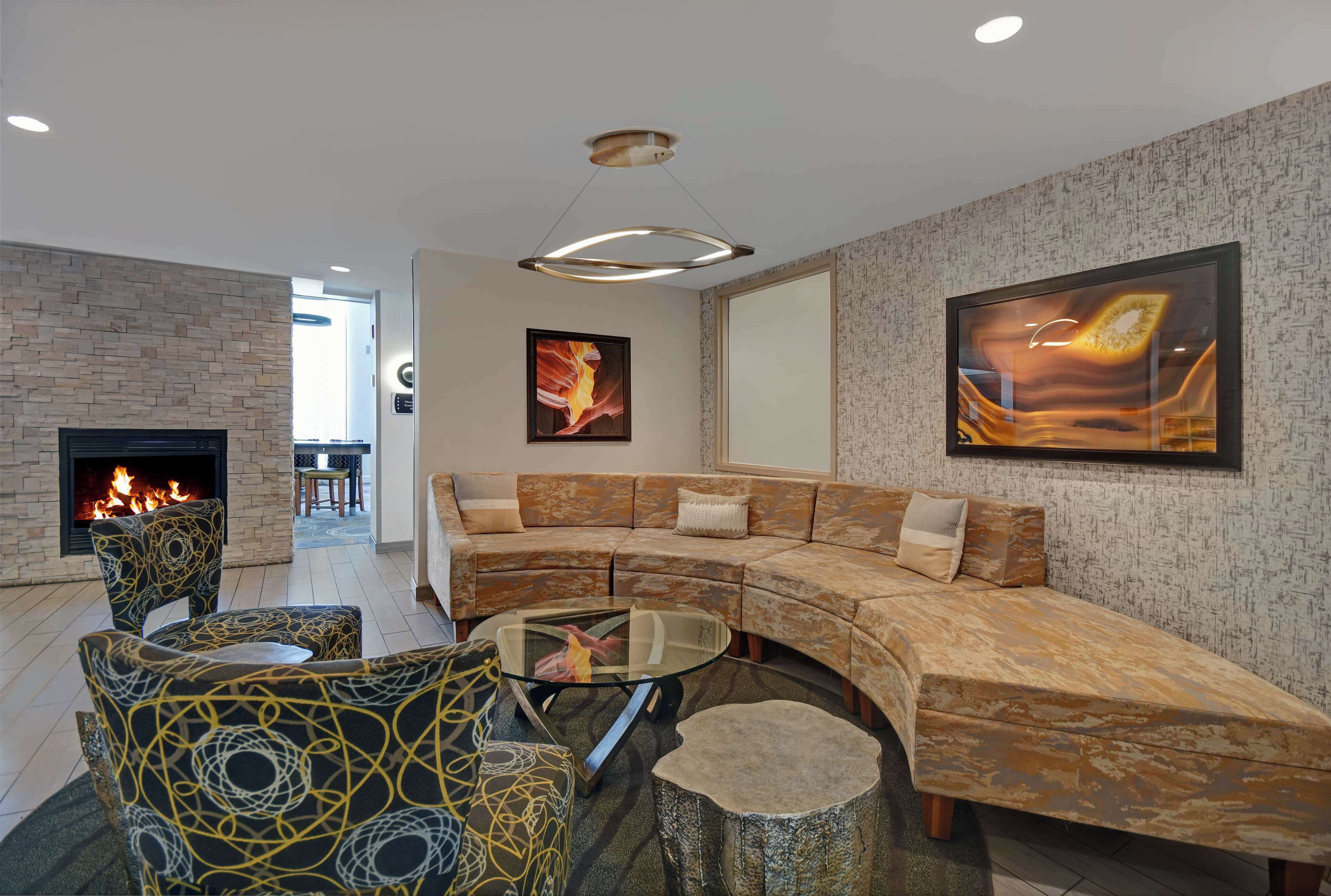 Una sala de estar moderna con chimenea de piedra, un sofá curvo, sillones estampados, una mesa de centro de vidrio y un cuadro abstracto de gran tamaño.