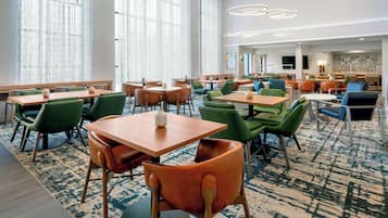 Un vestíbulo de hotel con varias zonas de estar, incluidas mesas y sillas, y ventanas grandes con cortinas translúcidas.