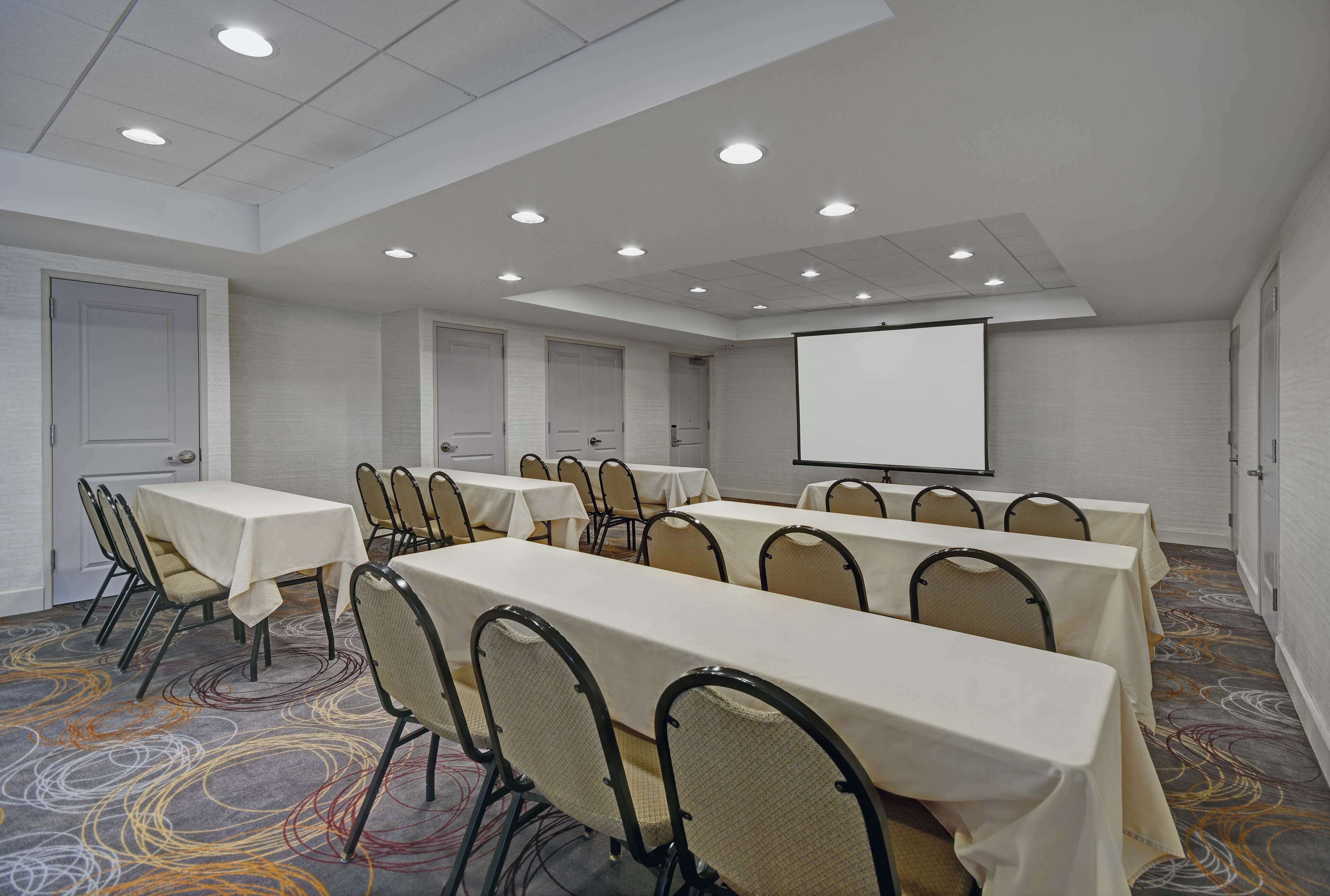 A conference room with multiple tables and chairs, a projector screen, and a carpeted floor.