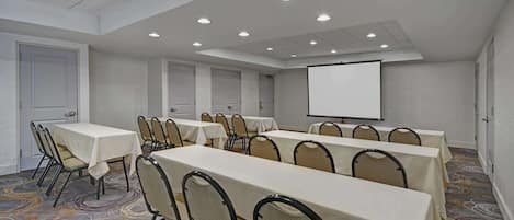 Una sala de conferencias con varias mesas y sillas, una pantalla de proyector y suelo alfombrado.