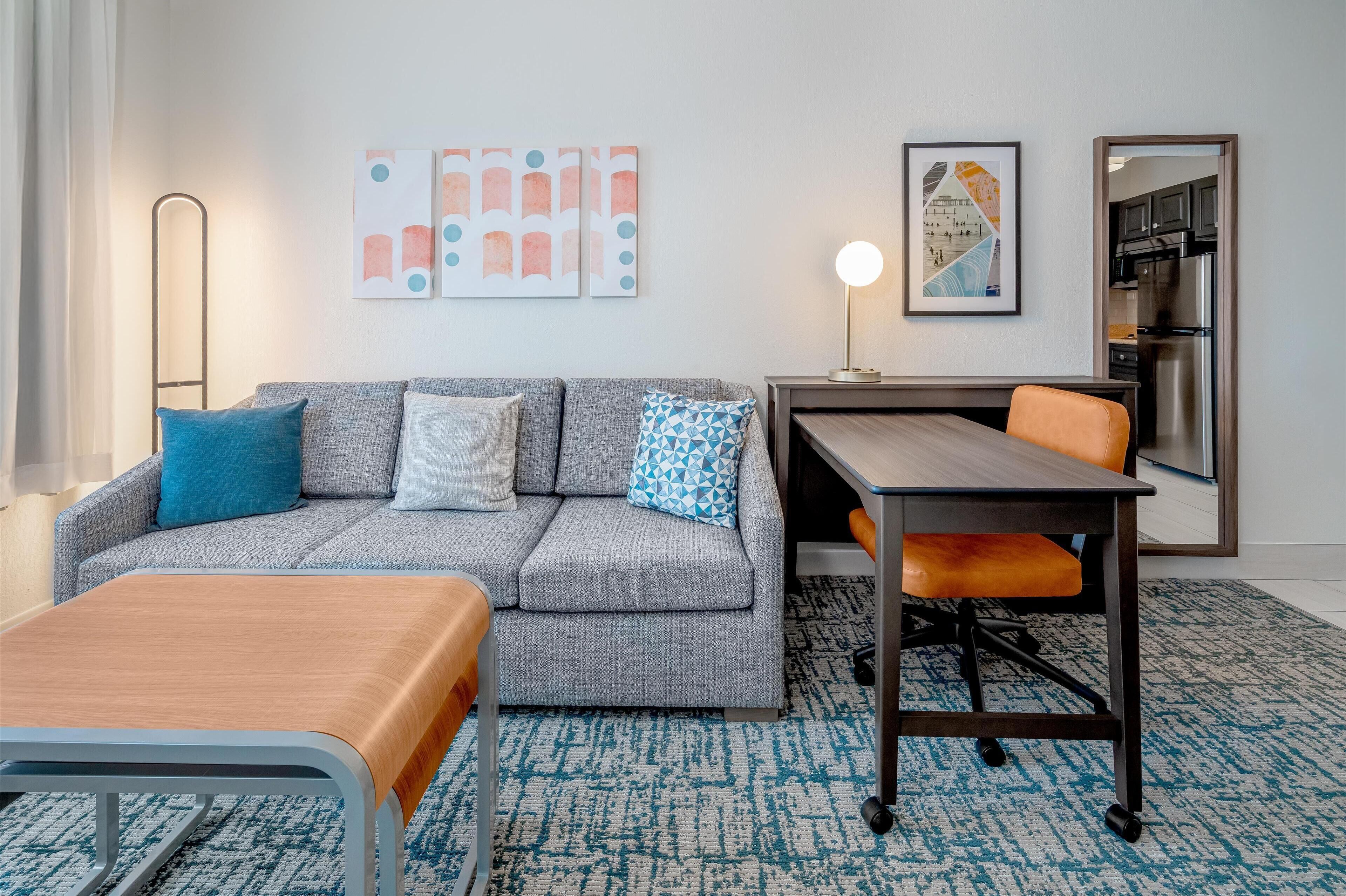 Una habitación de hotel moderna con un sofá gris, una mesa de madera y una alfombra azul estampada.