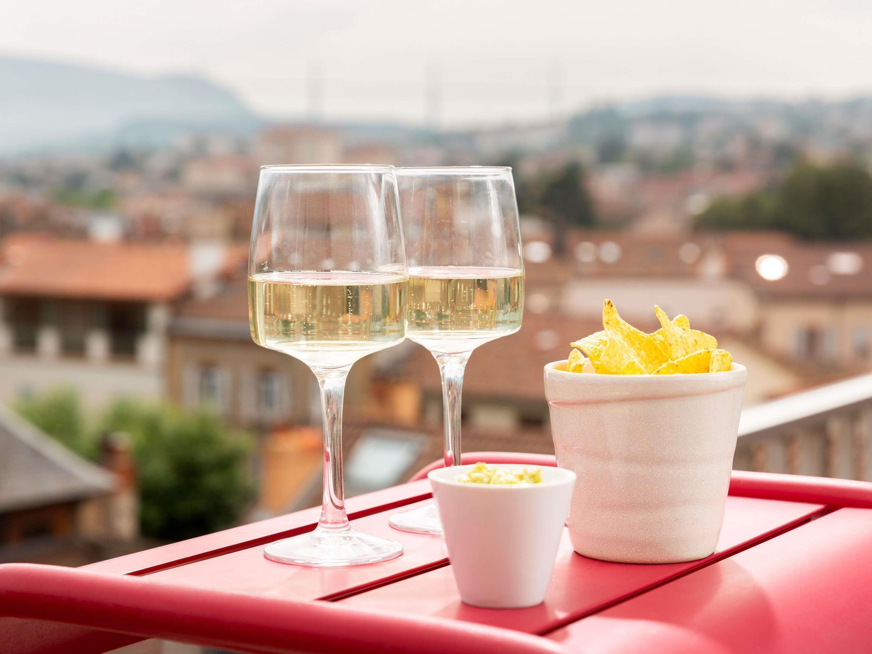 Dos copas de vino blanco sobre una superficie roja, junto a un cuenco de patatas fritas y un recipiente pequeño que parece ser una salsa o guarnición.