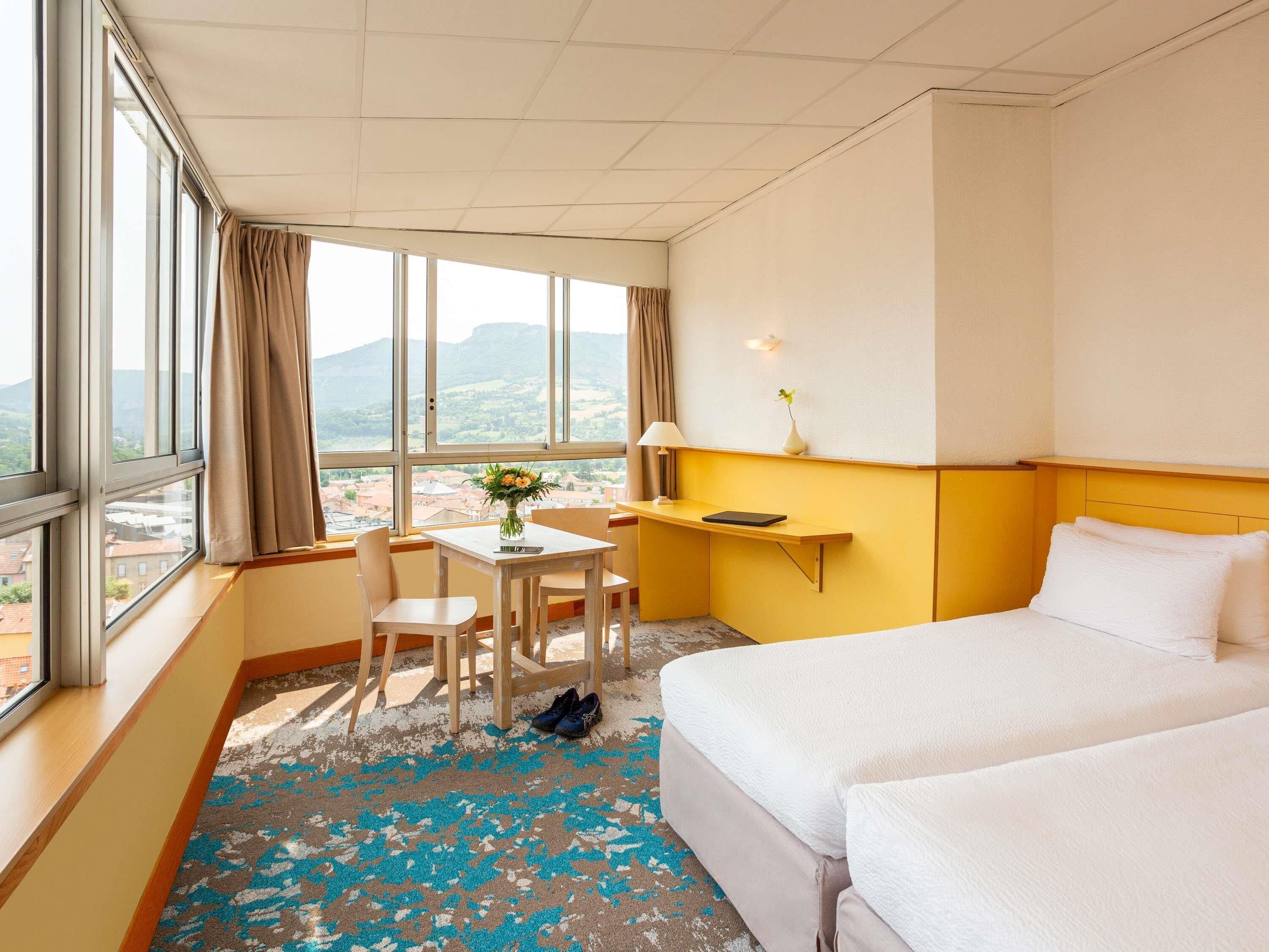 A hotel room with a bed, a desk, a chair, a lamp, and a view of the city.