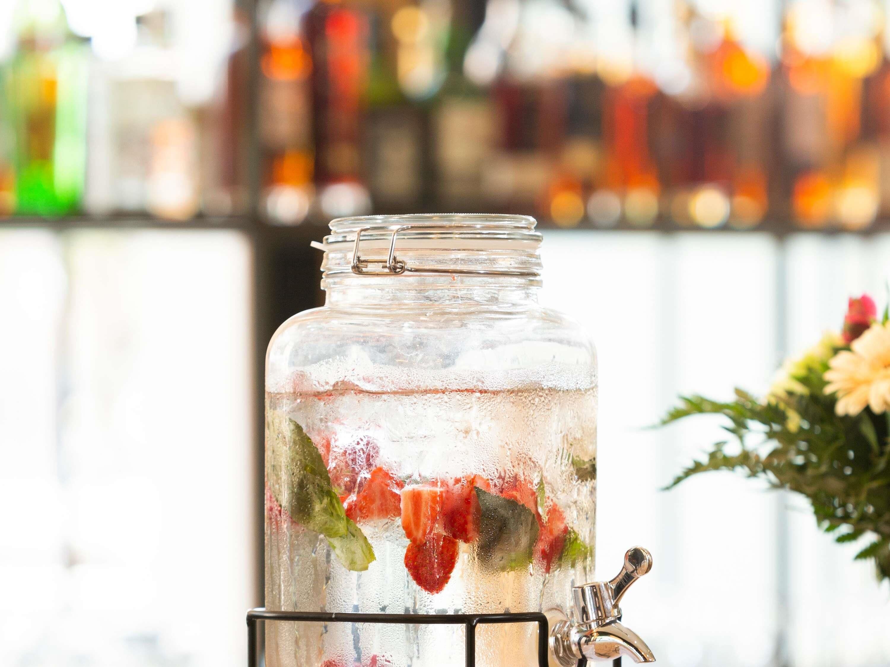 A glass jar with a metal tap, containing a beverage with ice and fruit pieces, set against a blurred background of various bottles.