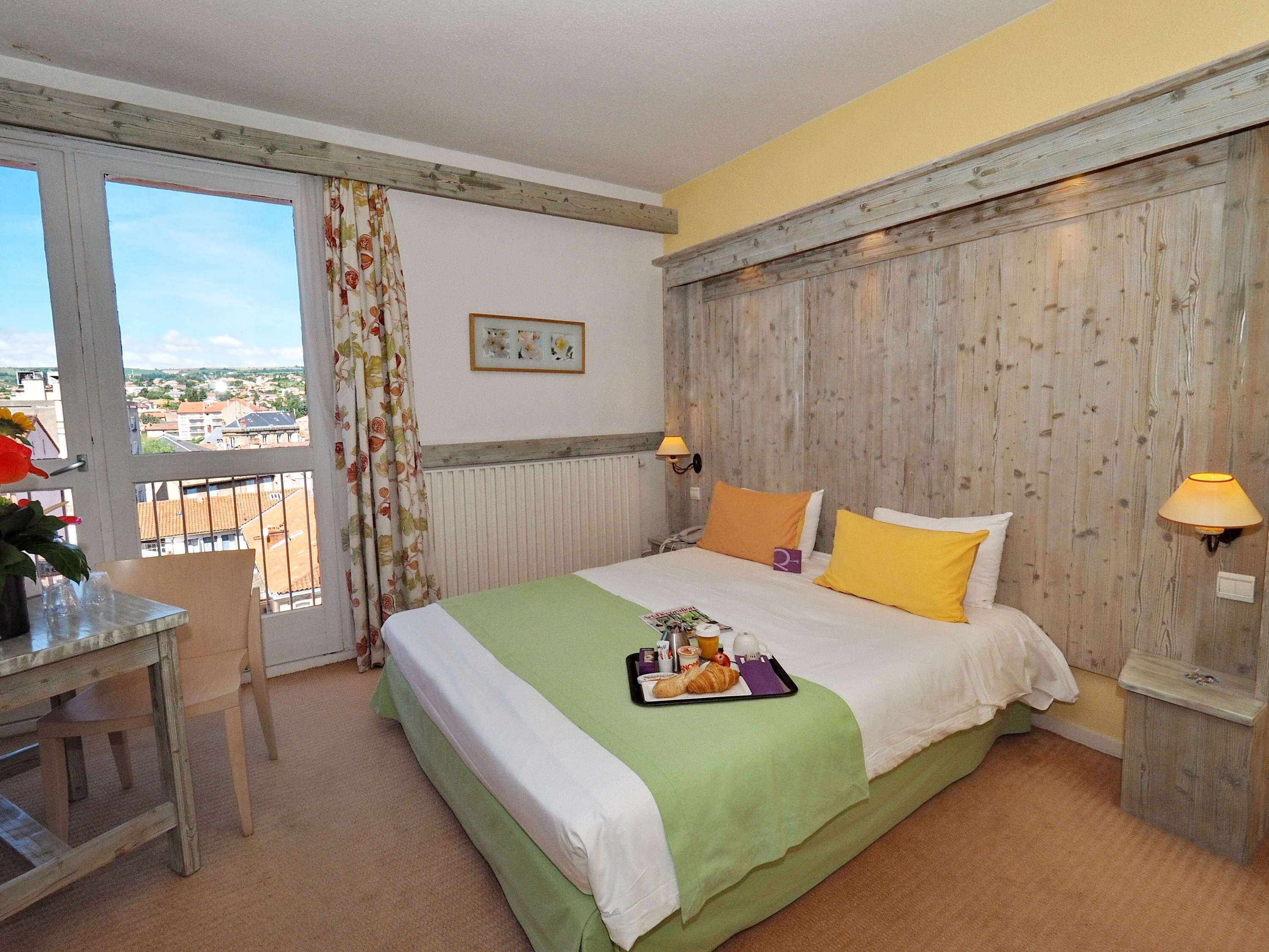 Habitación de hotel con cama, escritorio, silla, balcón y vistas, además de un cabecero de madera.