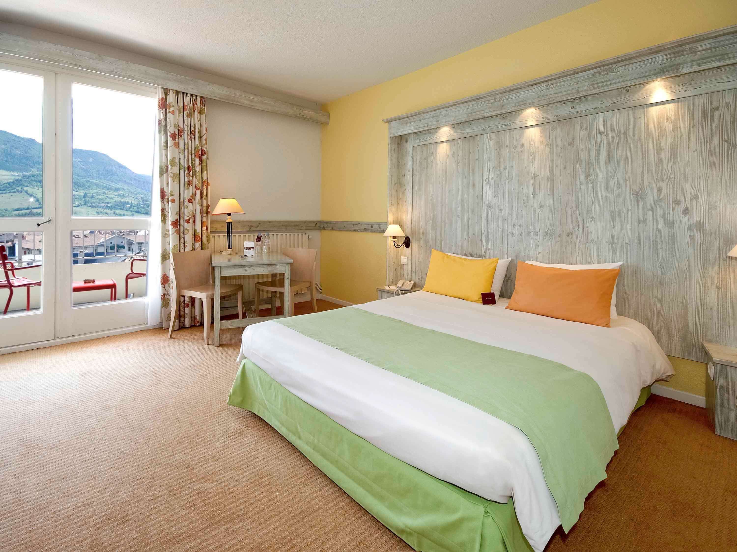 A hotel room with a large bed, a desk with a chair, a small table, and a view of the outdoors through a sliding door.