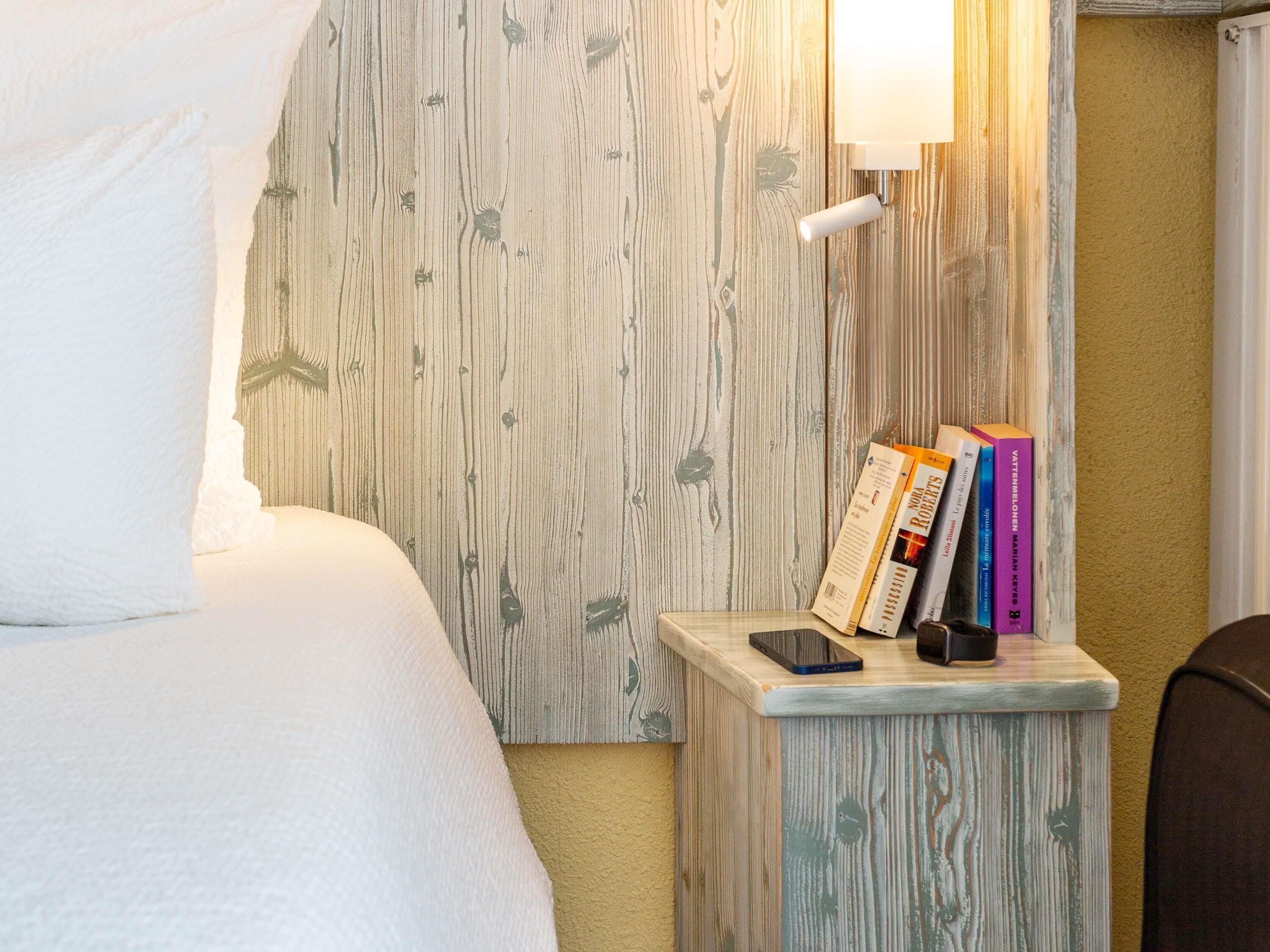 A neatly made bed with a pillow, a bedside table with a lamp, and a stack of books on the wall.
