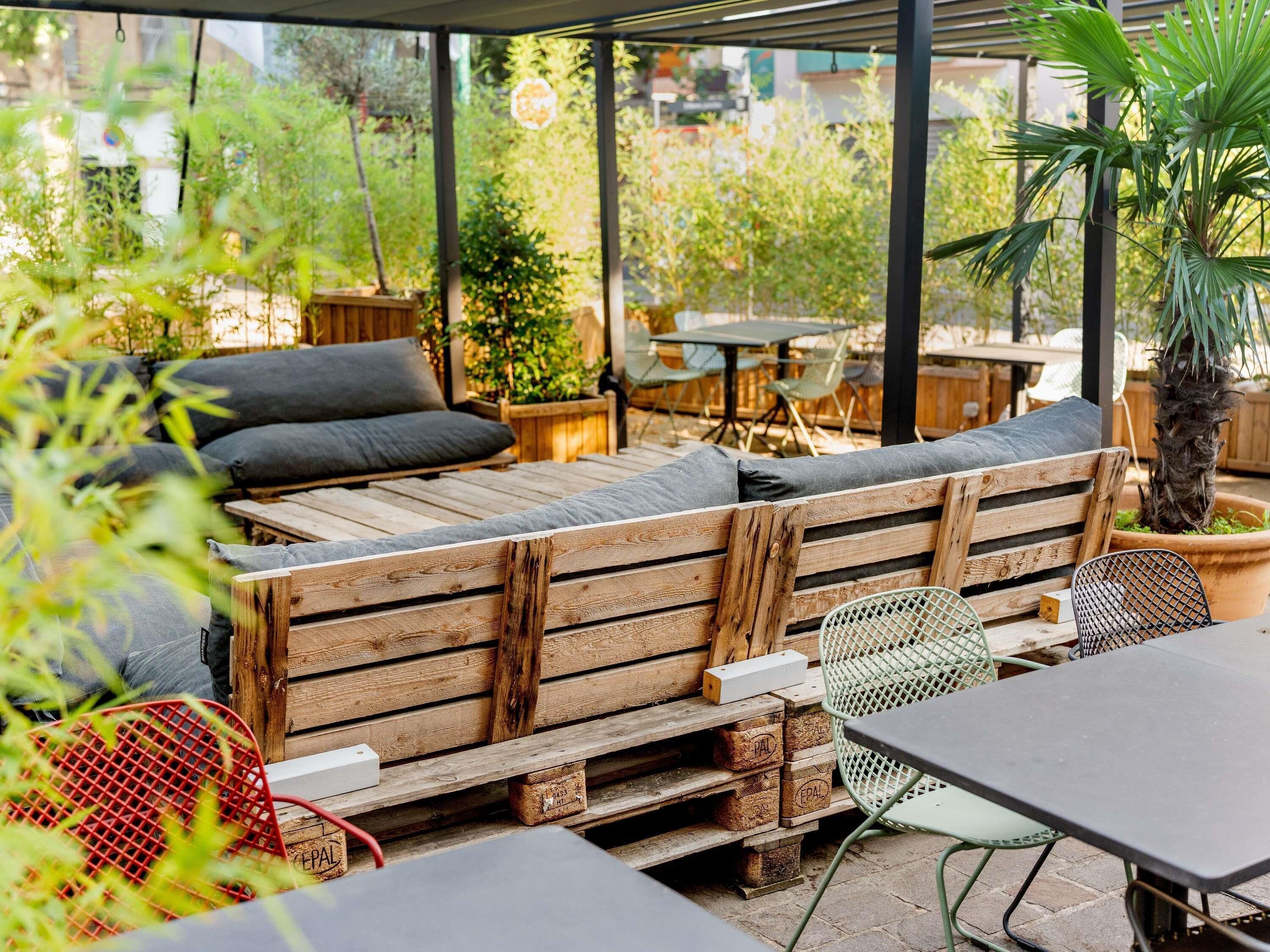Un espacio al aire libre con bancos de madera y sillas de metal, rodeado de vegetación y vistas a la ciudad.