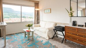 A hotel room with a white sofa, a small table with a vase of flowers, a dresser, and a large window offering a view of a cityscape and mountains. - Mercure Millau (Millau)