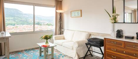 Habitación de hotel con un sofá blanco, una mesita con un florero, un aparador y un ventanal con vistas a un paisaje urbano y montañas.