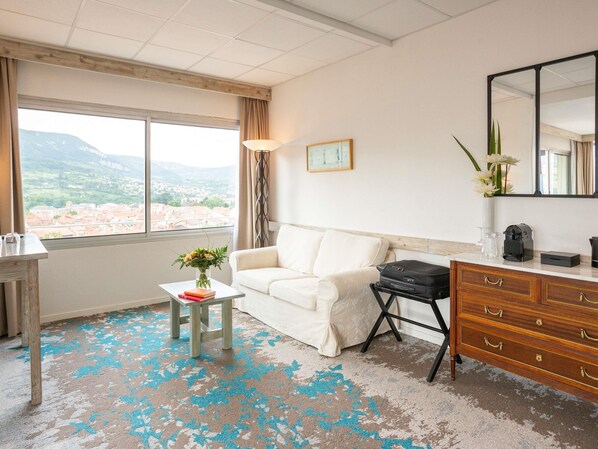 A hotel room with a white sofa, a small table with a vase of flowers, a dresser, and a large window offering a view of a cityscape and mountains. - Mercure Millau (Millau)