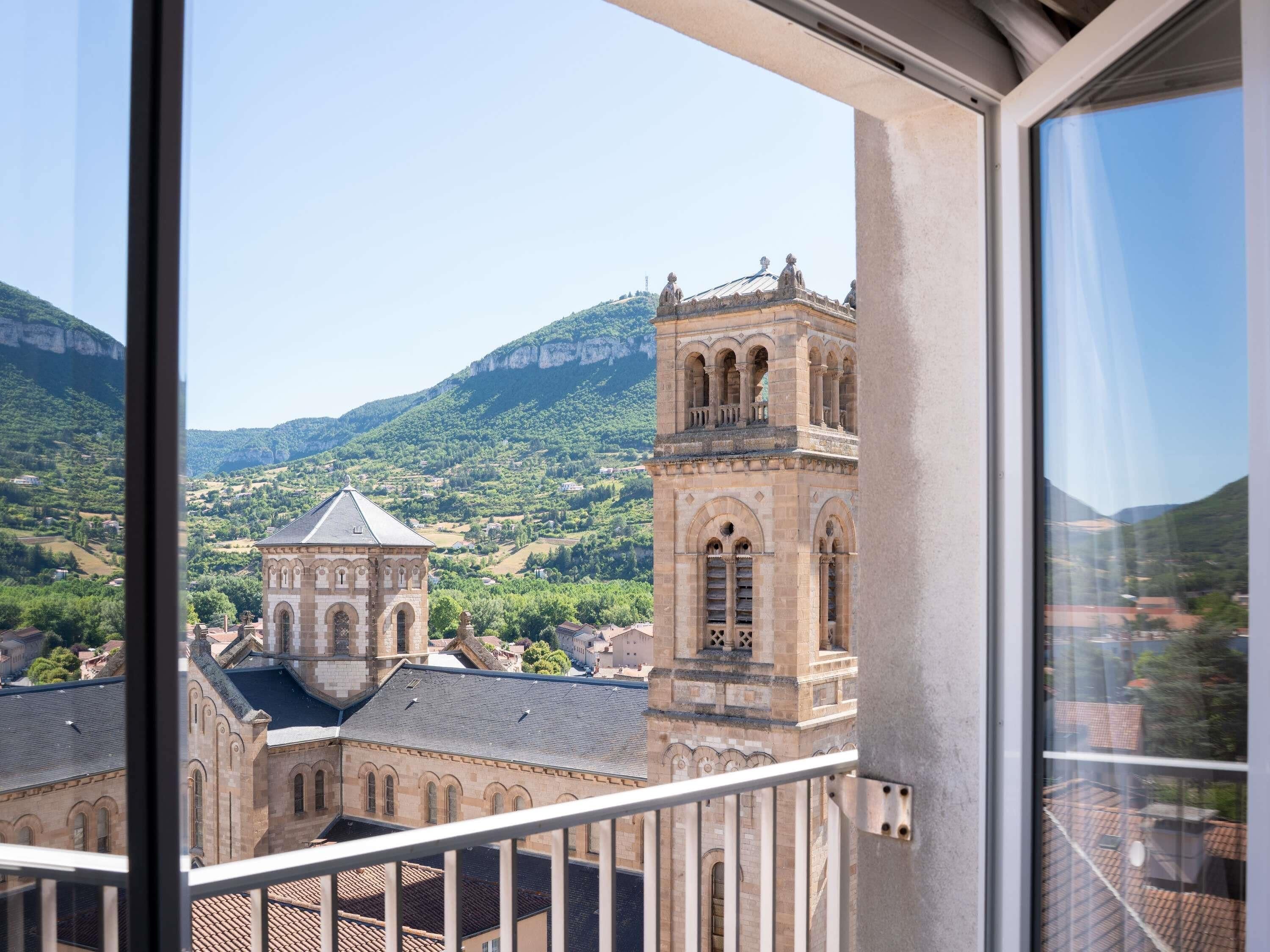 Blick von einem Balkon auf ein historisches Gebäude mit Glockenturm, vor der Kulisse von grünen Hügeln und einem klaren Himmel.