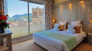A hotel room with a large bed, two bedside lamps, a view of a historic building, and a balcony with a railing. - Mercure Millau (Millau)