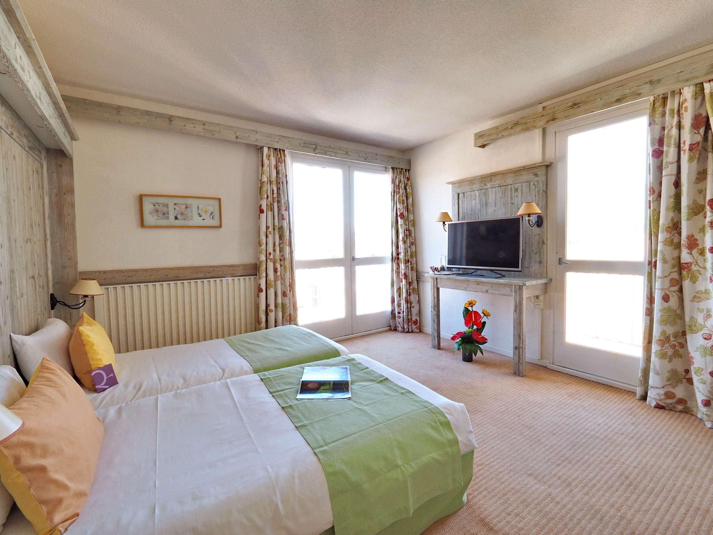 A hotel room with a bed, a television on a wooden desk, and large windows with floral curtains.