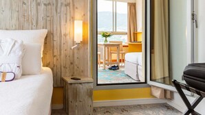 A hotel room with a bed, a small bedside table, a lamp, a window with a view, and a door leading to another room. - Mercure Millau (Millau)