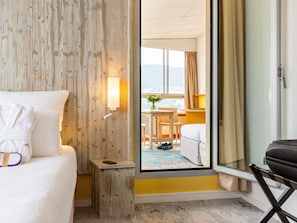 Una habitación de hotel con cama, una mesita de noche pequeña, una lámpara, una ventana con vistas y una puerta que da a otra habitación.
