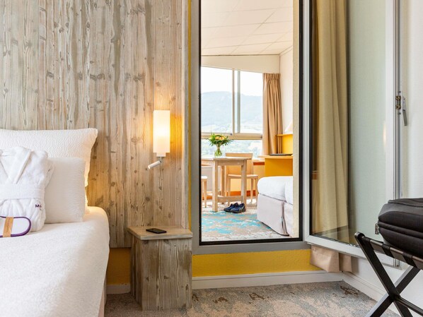Quarto de hotel com cama, uma pequena mesa de cabeceira, abajur, janela com vista e porta que dá para outro quarto.
