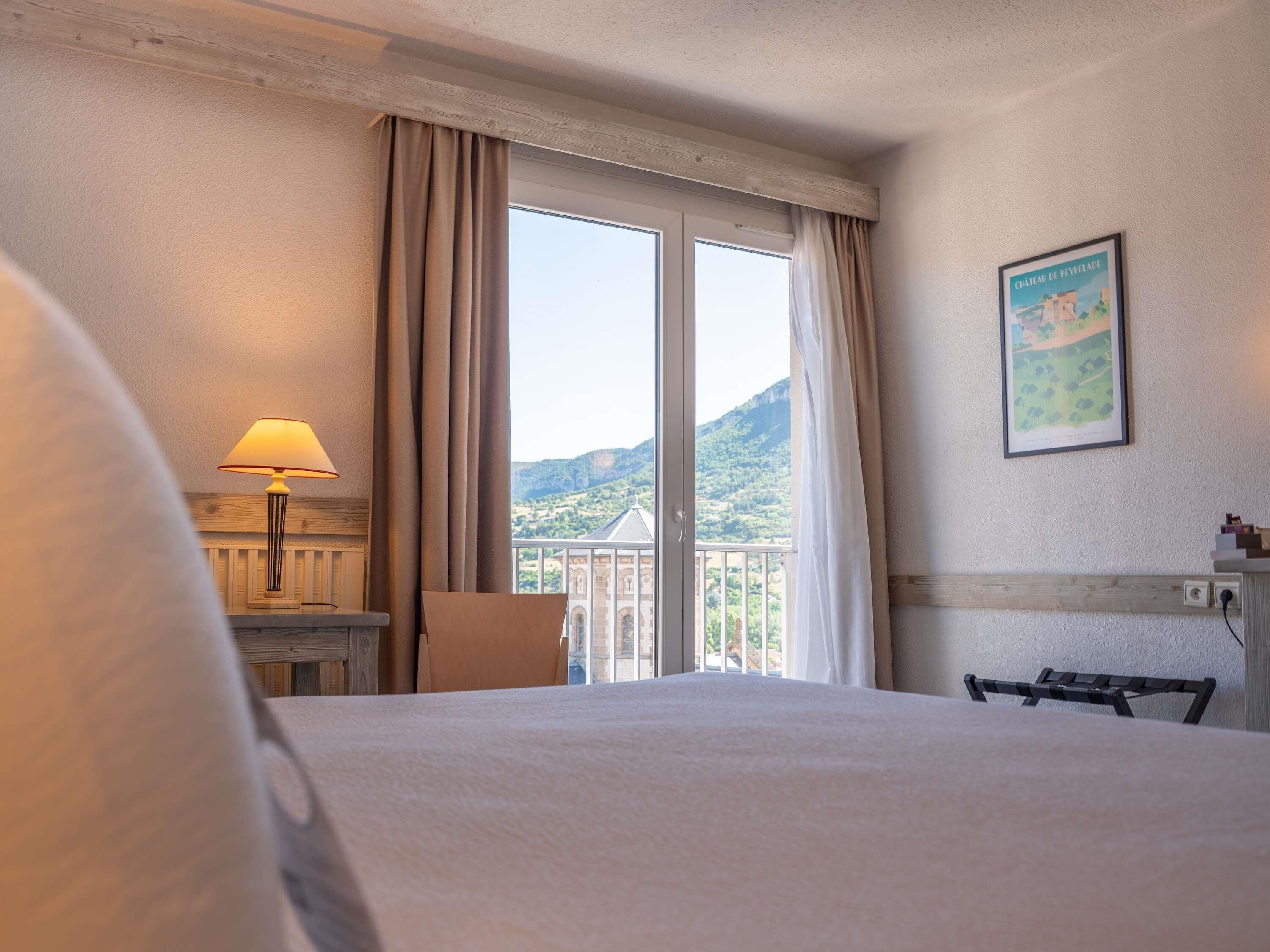 Une chambre d’hôtel avec un lit, un bureau, une chaise, une lampe et une grande fenêtre avec des rideaux.