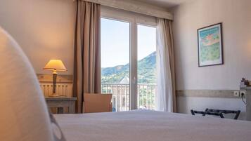 Une chambre d’hôtel avec un lit, un bureau, une chaise, une lampe et une grande fenêtre avec des rideaux.