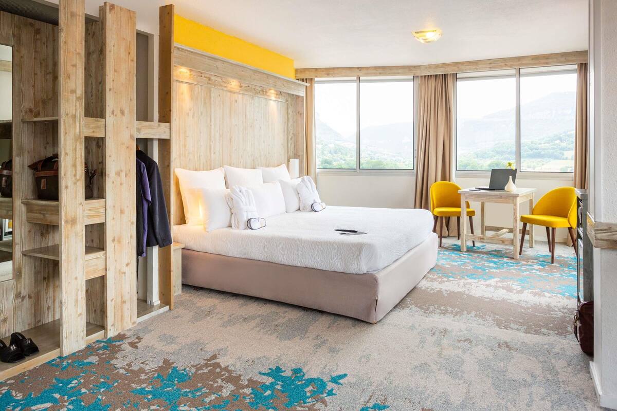 A hotel room with a bed, a desk, two chairs, a wooden wardrobe, and a view of the outdoors.