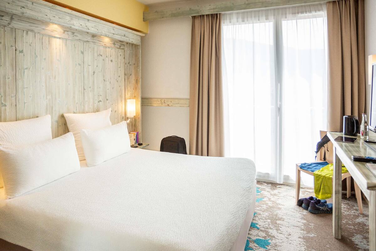 A hotel room with a large bed, a desk, a chair, and a window with curtains.