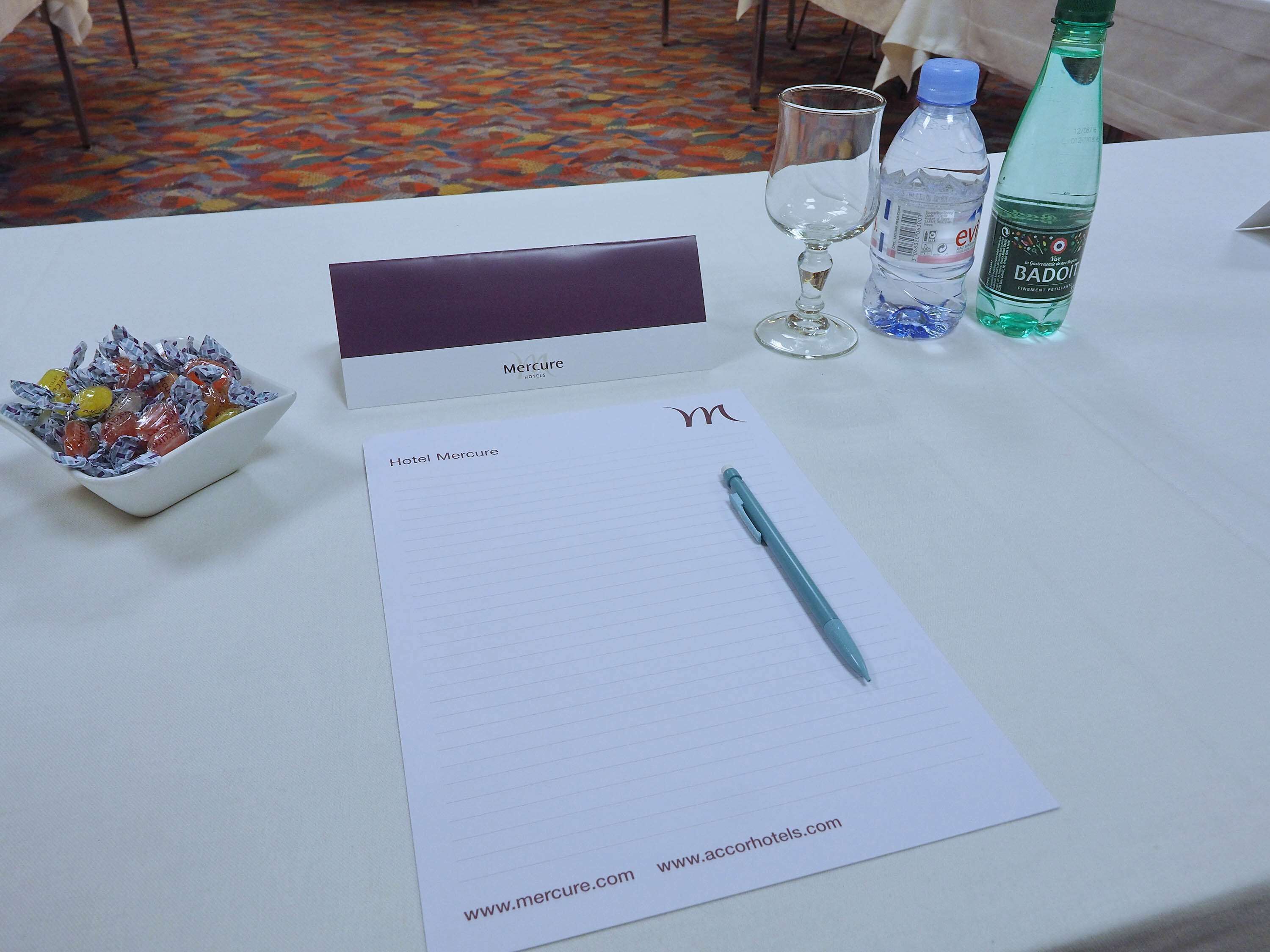 A hotel name card, a glass, a bottle of water, a bowl of candies, and a notepad with a pen on a table.