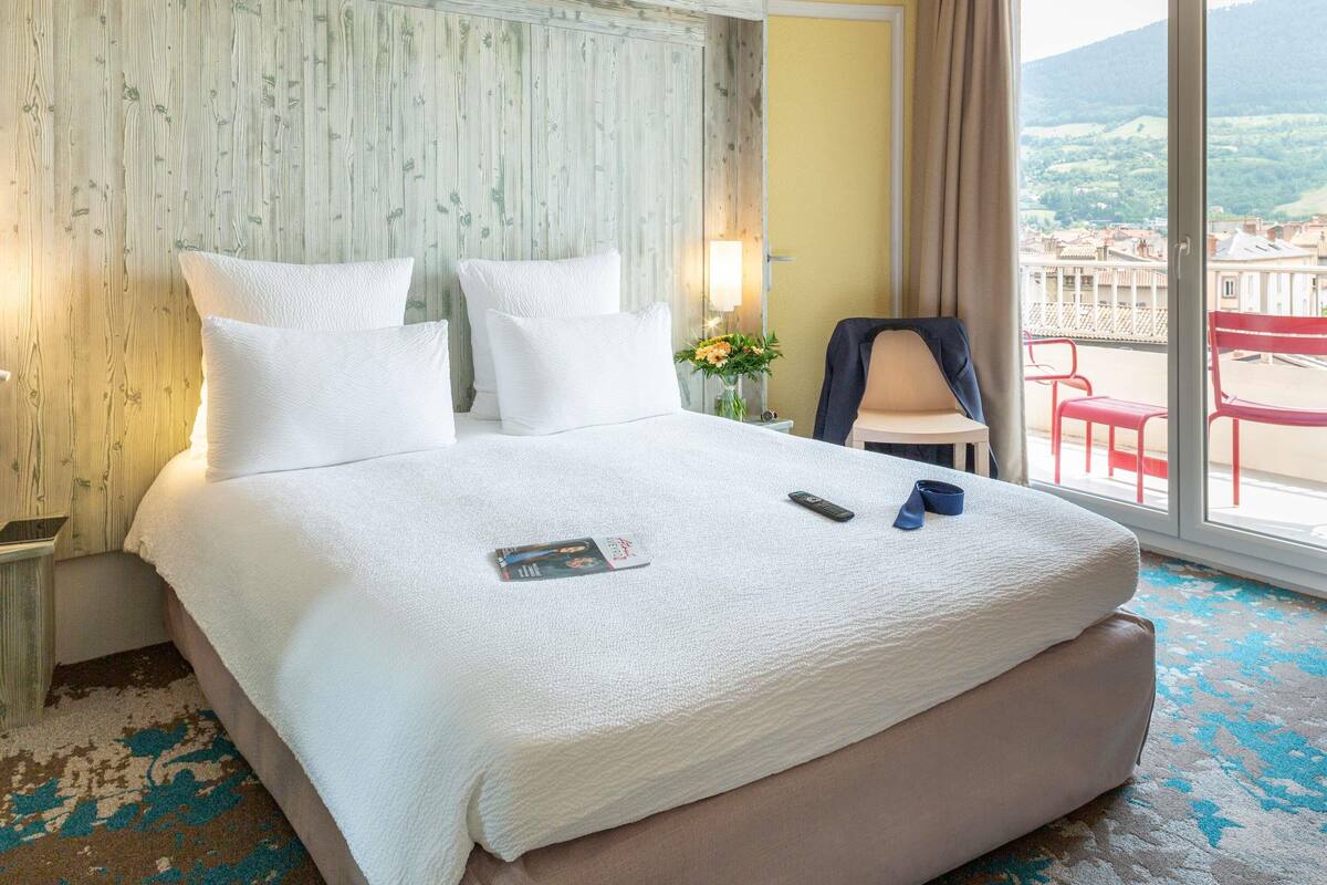A hotel room with a large bed, a chair, a table, and a view of the city.