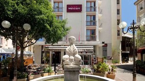 A fountain with a statue in the center, surrounded by potted plants. In the background, there is a building with a sign that reads "Mercure." - Mercure Millau (Millau)