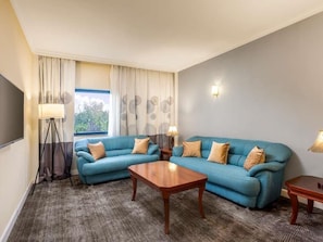 Habitación de hotel con sofá azul, una mesa de centro de madera, lámpara y vistas al exterior.